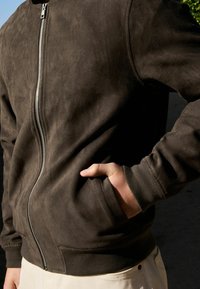 Chaqueta marrón con cremallera, hecha de un material similar al ante. Con puños acanalados, bolsillos laterales y una textura suave.