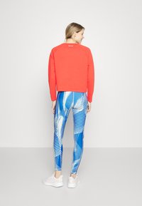 Felpa rossa corta con maniche lunghe, abbinata a leggings blu con motivi geometrici bianchi e strisce. Scarpe da ginnastica bianche.