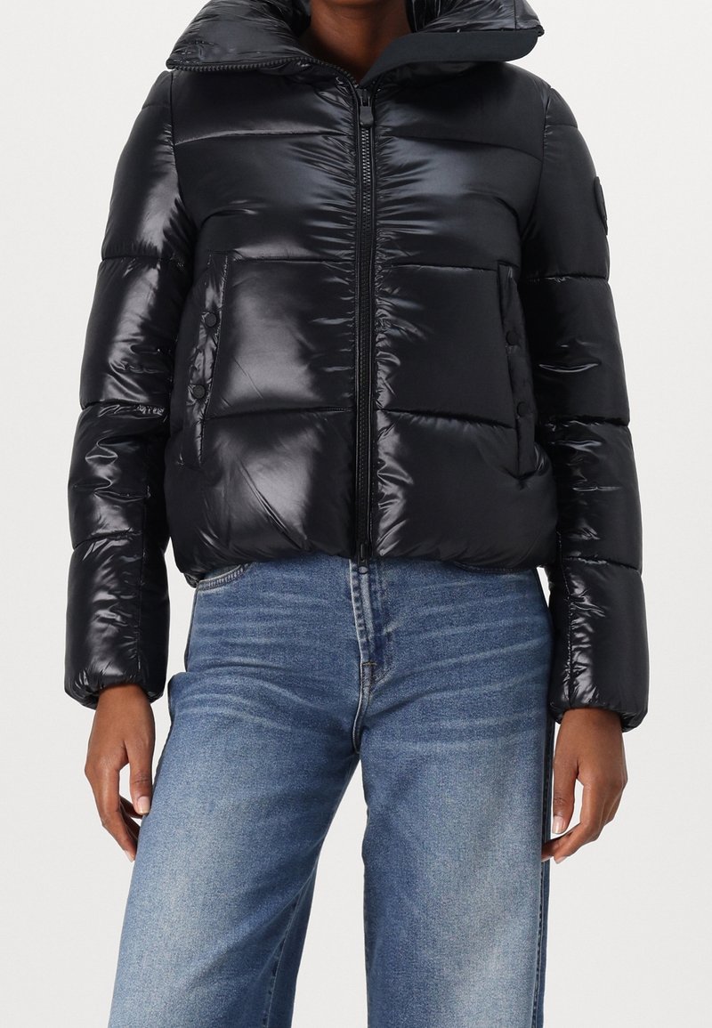 Doudoune noire brillante avec fermeture éclair à l'avant et poches à boutons-pression portée avec un jean bleu de teinte moyenne, coupé à la taille.