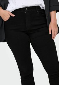 Fekete skinny farmernadrág sima textúrával, elülső zsebekkel és elől gombos zárással. Világos színű toppal viselve.