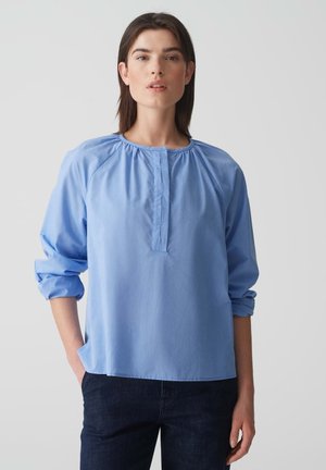 Lichtblauwe blouse met lange mouwen, met geplooide halslijn en zachte stof. Heeft een knoopsluiting en een comfortabele pasvorm. Gecombineerd met donkere jeans.