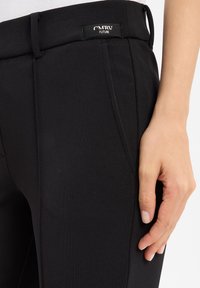 Schwarze Hose aus strukturiertem Stoff mit einem subtilen Muster. Verfügt über ein Etikett am Bund, schräge Taschen und eine taillierte Passform.