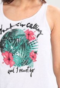 Witte tanktop met een cirkelvormige grafiek van tropische bladeren en rode hibiscus bloemen, met de tekst "Avontuur roept en ik moet gaan."
