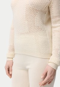 Hellbeiger Strickpullover mit verschiedenen Texturen und Mustern, mit einem gerippten Saum und Bündchen. Kombiniert mit weichen beigen Hosen.