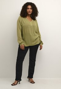 Blusa verde oliva a maniche lunghe con scollatura a V, tessuto testurizzato e vestibilità rilassata, abbinata a pantaloni neri slim-fit e tacchi neri.