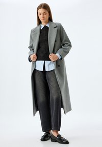 Grå uldblandingsfrakke med dobbeltradet front, lagdelt over en sort kabelstrik sweater og lyseblå skjorte, parret med mørke jeans og loafers.
