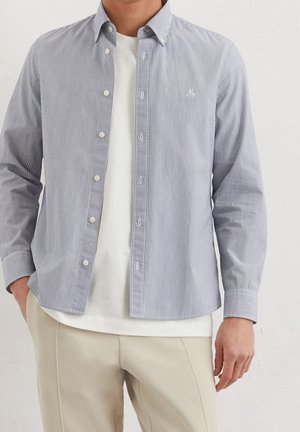 Mand iført beige bukser, hvid t-shirt og åben lys blå og hvid tyndstribet skjorte med lange ærmer mod en ensfarvet væg.