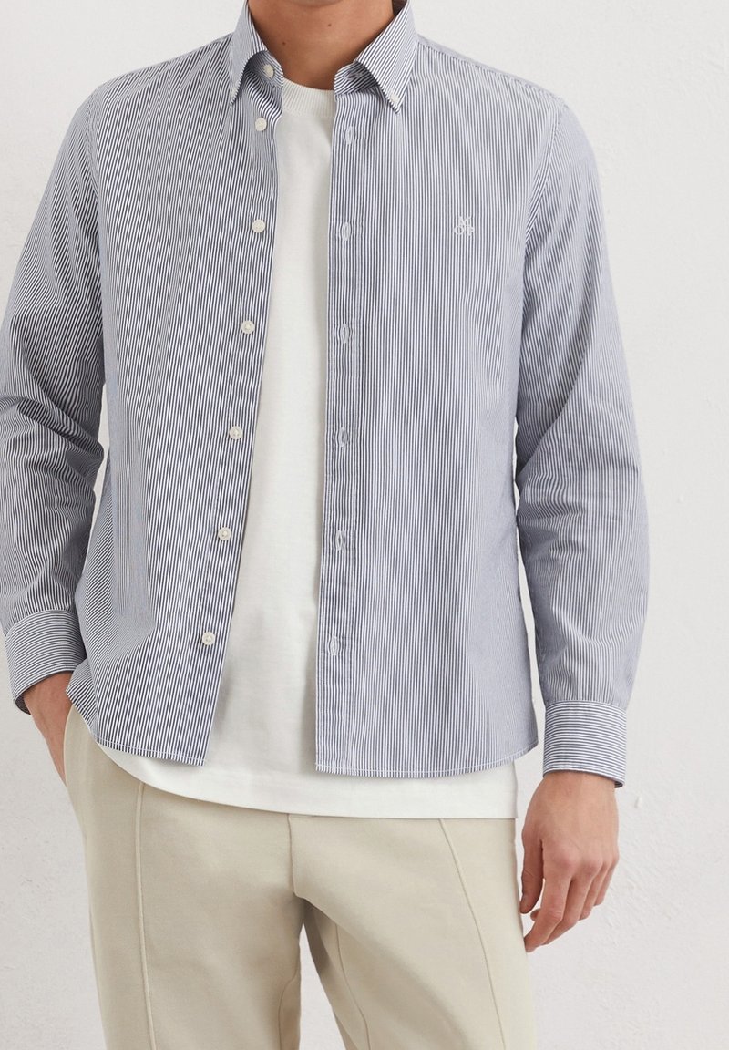 Homme portant un pantalon beige, un t-shirt blanc et une chemise à fines rayures bleu clair et blanc à manches longues, déboutonnée, contre un mur uni.