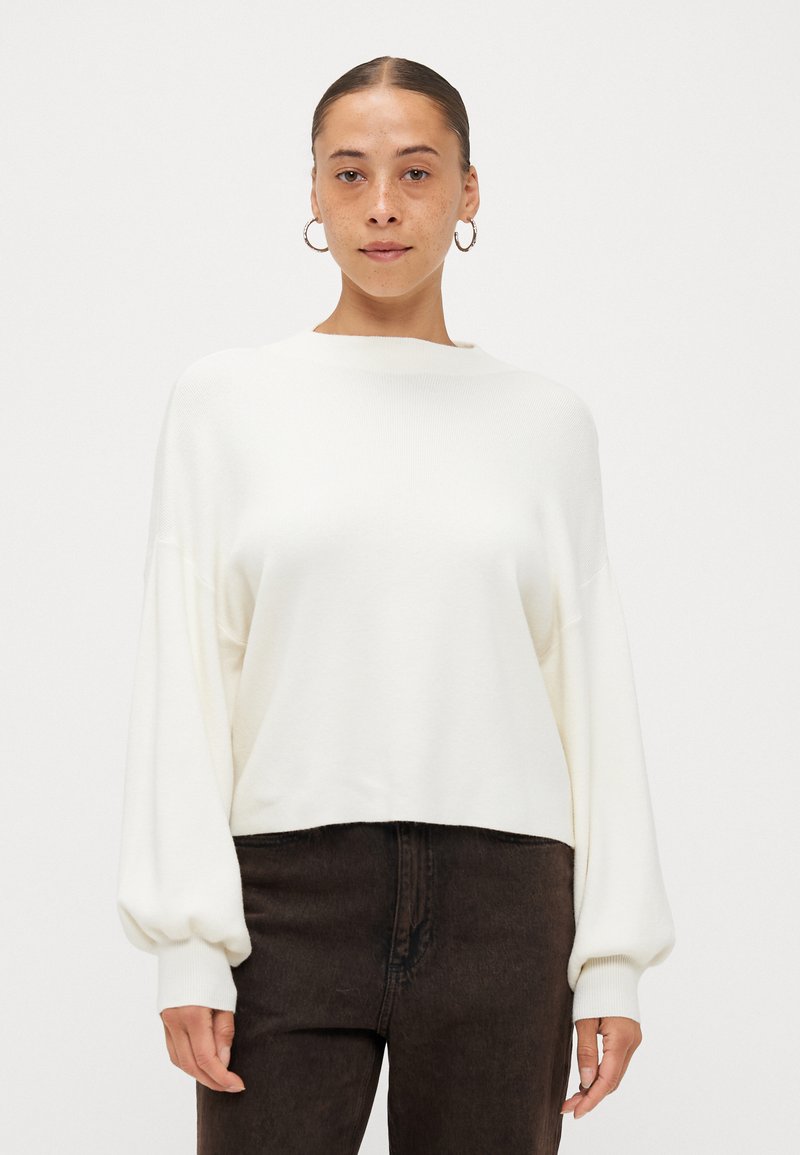 Weißer, kurzer Pullover mit langen, leicht puffigen Ärmeln und einem Boots-Ausschnitt, kombiniert mit dunkelbraunen Jeans. Weicher Stoff, minimalistisches Design.