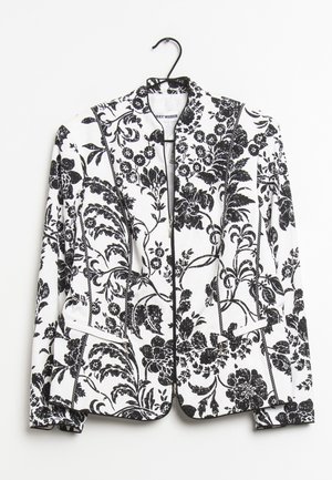 Blazer blanc avec motif floral et feuilles noires, coupe structurée, manches longues et poches avant, suspendu sur un cintre noir.