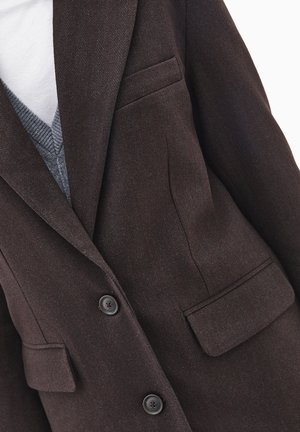 Brauner Fischgrätblazer mit zwei vorderen Knöpfen, zwei Seitentaschen und einem Reverskragen. Über einem grauen V-Ausschnitt-Pullover getragen.