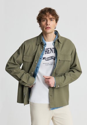 Giovane uomo con capelli ricci che si allaccia una giacca verde sopra una camicia di jeans e una maglietta bianca con stampa grafica, in piedi davanti a uno sfondo chiaro e uniforme.