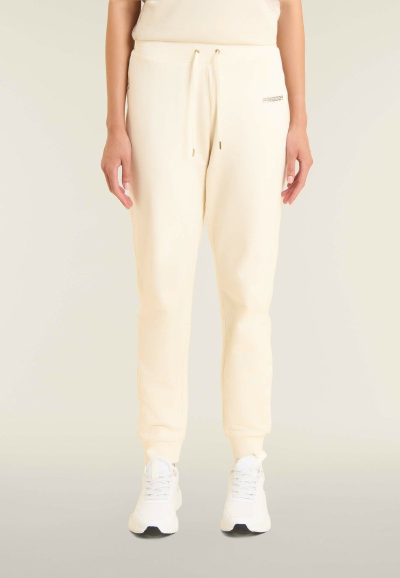 Pantalones de chándal color crema con textura acanalada, cintura elástica y detalle de cordón. Combinados con zapatillas deportivas blancas.