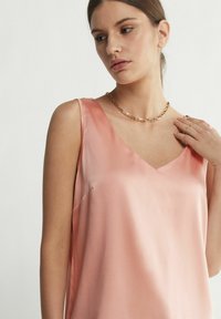 V-ringad, ärmlös topp i mjuk rosa satin, med en slät yta och subtil lyster. Modellen bär ett enkelt guldhalsband.