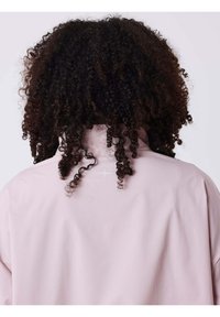 Chaqueta de manga larga de color rosa claro con cuello alto. Textura de tela lisa y detalle reflectante sutil en la parte trasera. Cabello rizado visible.