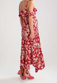 Rode maxi-jurk met bloemenprint, gerimpelde schouderbanden en een asymmetrische zoom. Voorzien van een roze-wit bloemenpatroon. Gecombineerd met roze schoenen met blokhak.