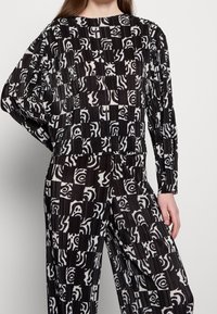 Czarny i biały kombinezon w wzory z plisowanym wykończeniem, z abstrakcyjnymi wzorami i luźnym krojem. Długie rękawy i okrągły dekolt.