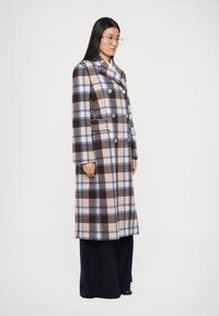 Manteau long à carreaux en laine mélangée beige, marron et bleu. Présente un design double boutonnage, de grands boutons et des poches latérales.