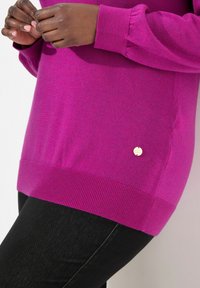 Fuchsia Pullover mit geripptem Saum und Bündchen, mit einem kleinen metallischen Logo-Akzent auf der unteren Seite, kombiniert mit schwarzen Leggings.