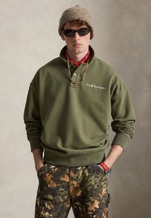 Νέος άνδρας με πράσινο φούτερ Polo Ralph Lauren, παντελόνι καμουφλάζ, πλεκτό καπέλο, γυαλιά ηλίου, πολύχρωμο κολιέ με χάντρες και κόκκινο βραχιόλι με ρολόι.