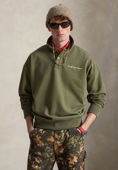 Νέος άνδρας με πράσινο φούτερ Polo Ralph Lauren, παντελόνι καμουφλάζ, πλεκτό καπέλο, γυαλιά ηλίου, πολύχρωμο κολιέ με χάντρες και κόκκινο βραχιόλι με ρολόι.