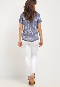 Modrá vzorovaná blůza z lehkého materiálu, s bílými geometrickými vzory a krátkým rukávem, v kombinaci s přiléhavými bílými džínami a béžovými podpatky.