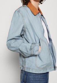 Ljusblå denimjacka med krage av brunt läder, sidofickor och dragkedjor. Har synlig sömnad och en avslappnad passform.