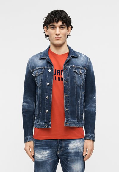 Jeune homme portant une veste en denim bleu délavée et un jean sur un t-shirt rouge avec motif, se tenant devant un fond clair uni.
