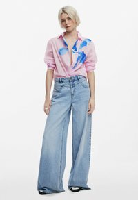 Camicia a maniche lunghe rosa chiaro con stampa astratta blu e scollatura a V profonda, abbinata a jeans in denim azzurro a gamba larga.