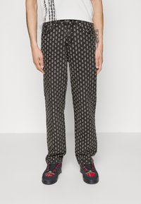 Musta farkut, joissa on vaaleanharmaa kuvioitu pintakuvio, viisi taskua, nappikiinnitys ja mustat ja punaiset kuvioidut kengät.