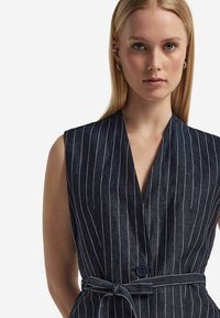 Ärmlös marinblå klänning med vita vertikala ränder, djup V-ringning och knäppning framtill. Inkluderar ett knytbälte i midjan för justering.