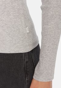 Maglia grigia a coste con maniche lunghe e etichetta del logo ben visibile, abbinata a pantaloni di jeans neri, che mostrano una texture morbida e un design aderente.