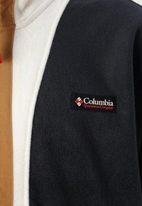 Giacca in pile a tre pannelli con sezioni di tessuto nero, bianco e marrone. Include una toppa con il logo della Columbia Sportswear Company.