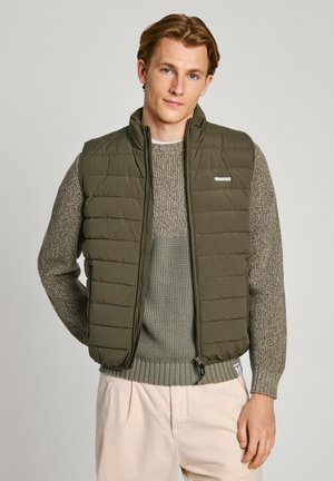 Chaleco acolchado de color verde oliva con textura de acolchado, cuello alto y cierre frontal de cremallera, usado sobre un suéter de punto gris y pantalones beige claros.