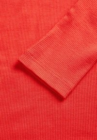 Czerwona tkanina koralowa z prążkowaną strukturą i gładką fakturą, z brzegiem rękawa wykończonym obszyciem oraz równomiernymi pionowymi liniami na całej powierzchni.