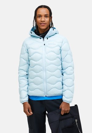 Light blue puffer jacket with a wavy quilted design, zip closure, hood, and branding. Worn over a black long-sleeve top.