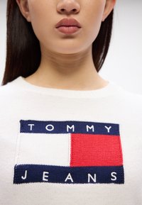Biely pletený sveter s obdĺžnikovým logom v modrej, červenej a bielej farbe a textom "TOMMY JEANS" v námorníckych písmenách.