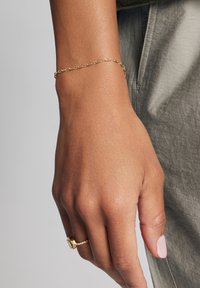 Złota bransoletka łańcuszkowa z powiązanym wzorem, prezentowana na dłoni o jasnej opaleniźnie. Bransoletka ma gładką teksturę i subtelny blask.