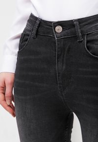 Los pantalones vaqueros negros de denim tienen una cintura alta, cinco bolsillos y un sutil efecto de desgaste. La tela tiene una textura suave con un ligero brillo.