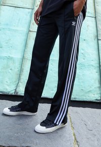 Zwarte sportbroek met drie witte stripes langs de zijkanten, gemaakt van gladde stof. Draag met zwarte sneakers met een witte zool.