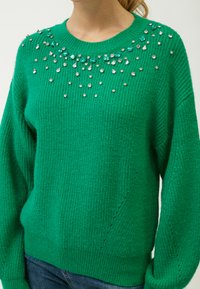 Grüner Strickpullover mit Rundhalsausschnitt, verziert mit klaren und smaragdgrünen Strasssteinen am Kragen. Lockere Passform, strukturiertes Material.