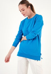 Sudadera azul de manga larga en una tela suave con puños y dobladillo acanalados. Presenta aberturas laterales para mayor comodidad y un ajuste holgado, combinada con pantalones crema.