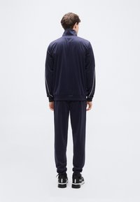 Ναυτικό φόρμα με ψηλό γιακά και λευκές ρίγες στα πλάγια, κατασκευασμένη από απαλ ό ύφασμα με χαλαρή εφαρμογή. Περιλαμβάνει παντελόνι που ταιριάζει και μαύρα αθλητικά παπούτσια.