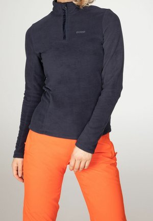 Frau, die einen schwarzen Fleece mit Reißverschluss und langen Ärmeln sowie leuchtend orange Hosen trägt, steht mit einer Hand nahe ihrer Hüfte.