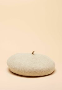 Bezs filckalapka, kerek formájú, puha textúrájú, apró cippentővel a tetején az állításhoz, letisztult dizájn, lágy barack színű háttér előtt.