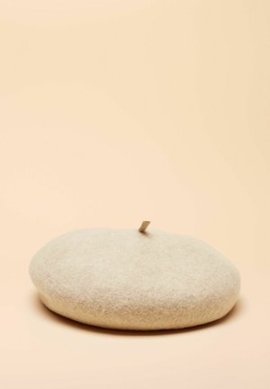 Beretă bej din fetru, formă rotundă, textură moale cu o mică clapetă în vârf pentru reglare, design minimalist pe un fundal roz pal.