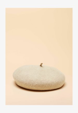 Beretă bej din fetru, formă rotundă, textură moale cu o mică clapetă în vârf pentru reglare, design minimalist pe un fundal roz pal.