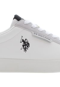 Bílé tenisky s hladkým povrchem a šedými akcenty. Obsahují logo U.S. Polo Assn. a potištěnou scénu s polokou na boku. Ploché tkaničky.
