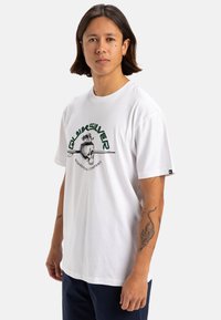 Camiseta de algodón blanca con cuello redondo, que presenta un gráfico verde de una calavera con palmeras y el texto "Quiksilver", de corte relajado.