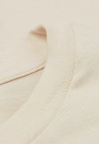 Světlý béžový bavlněný materiál s hladkou texturou a kulatým výstřihem. Švy jsou viditelné podél límce a lemu.